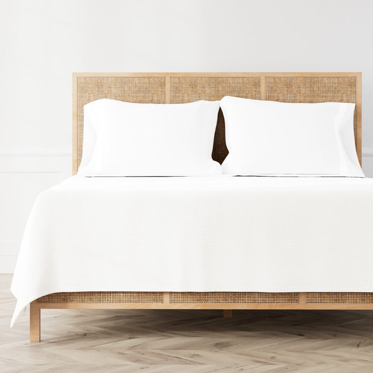 A neatly made bed featuring the Minaudiere Sheet Set in White‑Bone from the Judith Leiber Couture by Ann Gish collection, styled against a light wood rattan headboard and matching footboard in a bright, minimalist room. - SSMIK-WHI-BON SSMIQ-WHI-BON