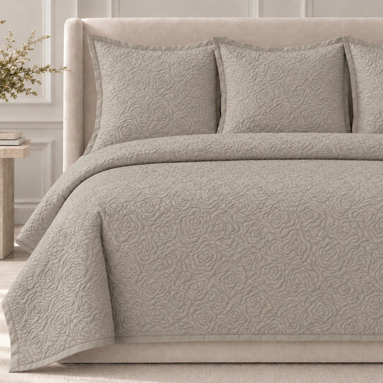 The Roses Coverlet Set from Judith Leiber Couture by Ann Gish, shown in Taupe, featuring its signature rose‑quilted pattern with matching shams, styled on a bed with a beige upholstered headboard in a softly lit, paneled bedroom. - YSETCORQK‑TAU YSETCORQQ‑TAU