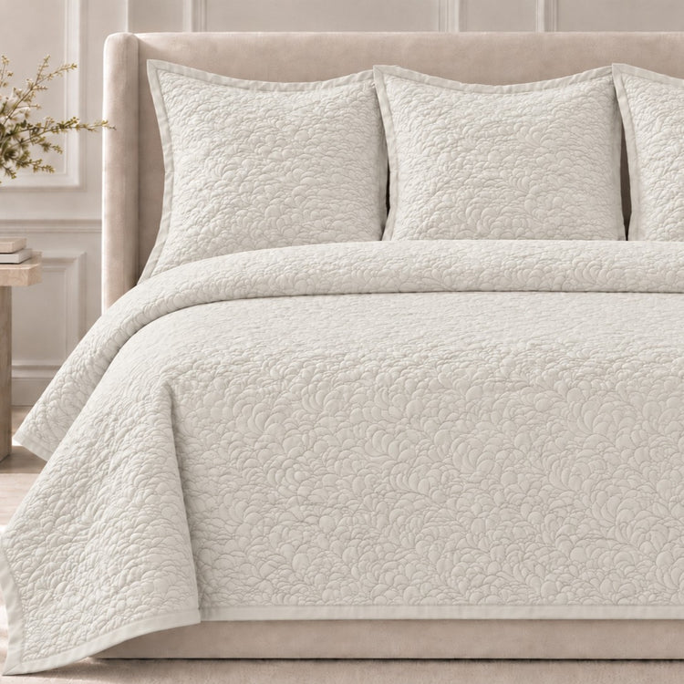 The Shells Coverlet Set from the Judith Leiber Couture by Ann Gish collection, shown in bone with its scalloped quilted pattern, styled on a bed with a beige upholstered headboard and matching shams in a softly lit bedroom. - YSETCOSHK-BON YSETCOSHQ-BON