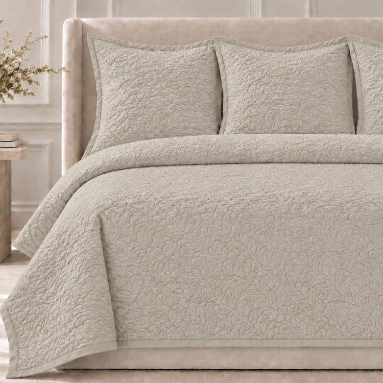 The Shells Coverlet Set from the Judith Leiber Couture by Ann Gish collection, shown in pumice with its scalloped quilted pattern, styled on a bed with a beige upholstered headboard and matching shams in a softly lit bedroom. - YSETCOSHK-PUM YSETCOSHQ-PUM