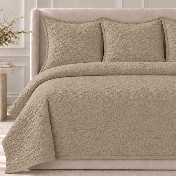 The Shells Coverlet Set from the Judith Leiber Couture by Ann Gish collection, shown in sand with its scalloped quilted pattern, styled on a bed with a beige upholstered headboard and matching shams in a softly lit bedroom. - YSETCOSHK-SAN YSETCOSHQ-SAN
